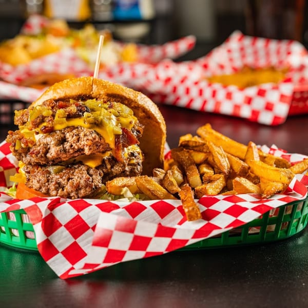 Double patty burger with cheese, bacon, and green chiles, served with fries in a green basket.