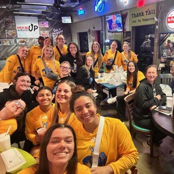 A large group of women in yellow shirts smiling and posing inside Mixed Up Burgers restaurant.