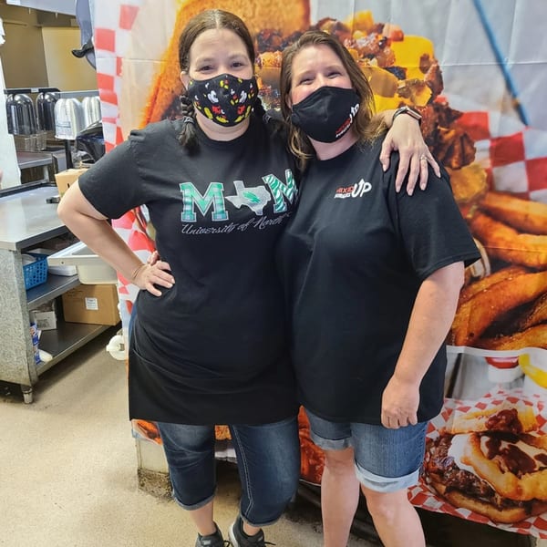 Two women in Mixed Up Burgers shirts and masks stand in front of a burger-themed backdrop.