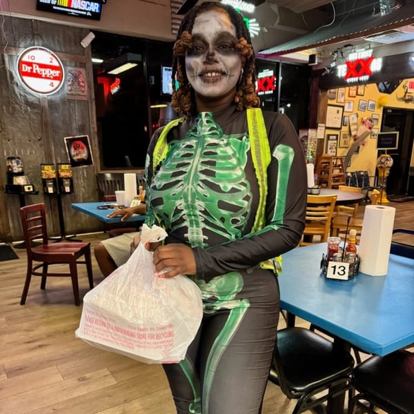 A person in a skeleton costume and face paint holds a takeout bag inside Mixed Up Burgers.