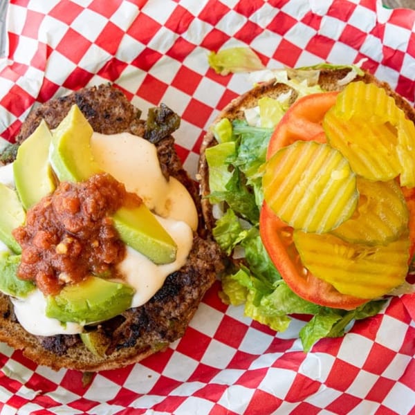 An open burger with avocado, salsa, and sauce on one half, and lettuce, tomato, and pickles on the other.