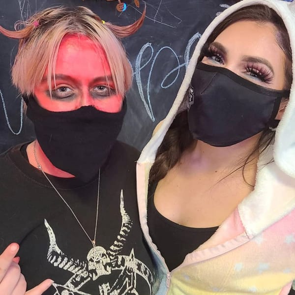 Two people in Halloween costumes and masks, one with red face paint and horns, against a chalkboard.